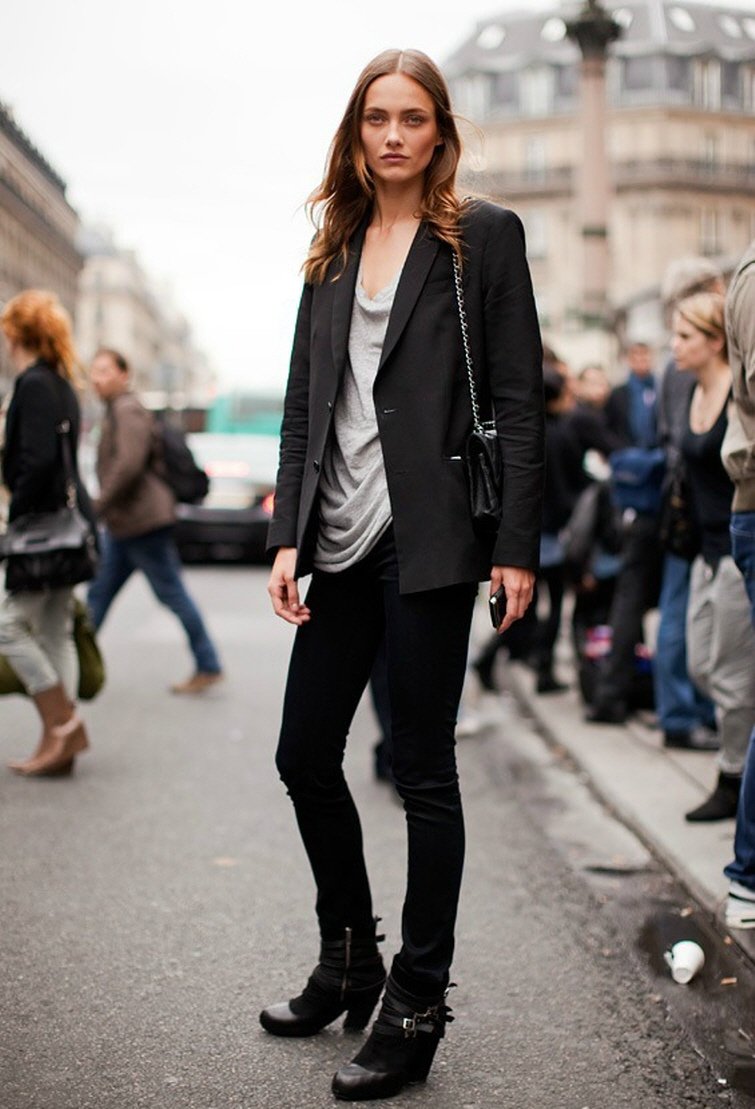 Modelo parada na rua tira foto de street style usando calça jeans preta, botas pretas, camiseta cinza, blazer preto e bolsa chanel preta