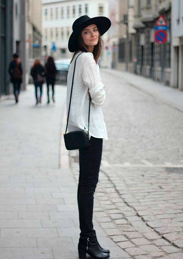 Mulher parada na calçada posa para foto usando calça jeans preta, bota preta, maxi tricot, bolsa preta a tiracolo e chapéu preto