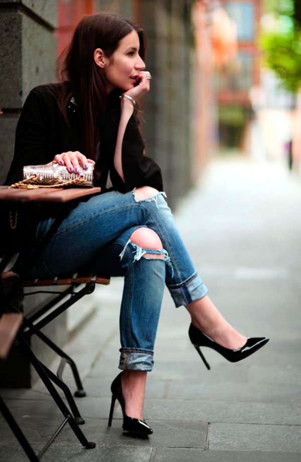 Irina Lakicevic sentada em mesa de café na rua posa para foto olhando para o lado usando distroyed jeans, scarpin preto, blazer preto e bolsa de pyton