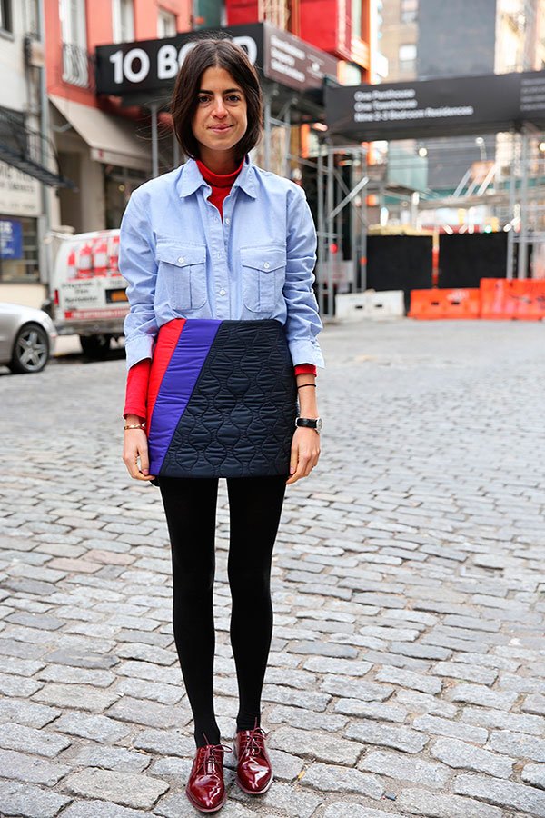 Leandra Medine posa para foto em meio às ruas de Nova York usando turtleneck vermelha, camisa de dois bolsos azul, saia de matelassê, meias pretas, oxford de verniz vinho