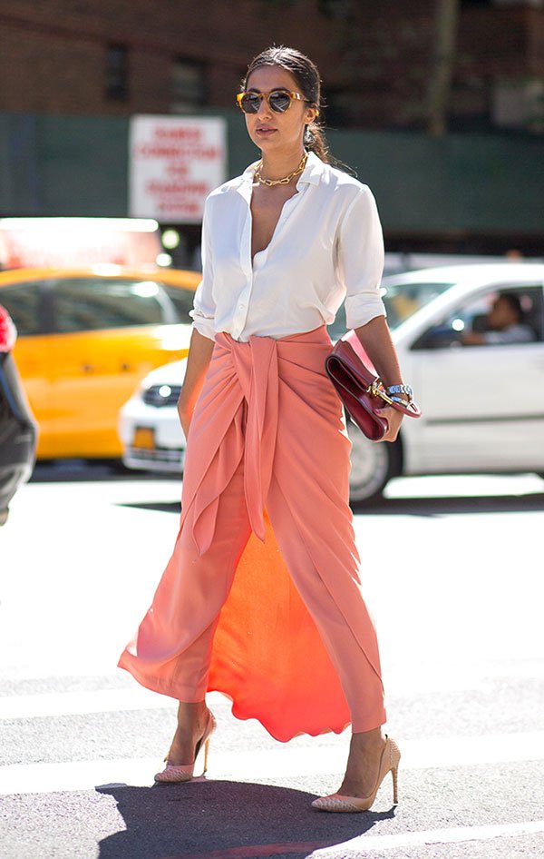 Mulher posa para foto no meio da rua com saia coral de amarração, camisa branca aberta e segurando uma clutch