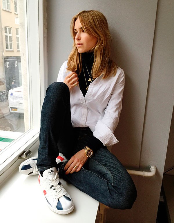 Pernille Teisbaek sentada em frente à uma janela, posa pra foto usando turtleneck preta, camisa branca, calça jeans e tênis stan smith
