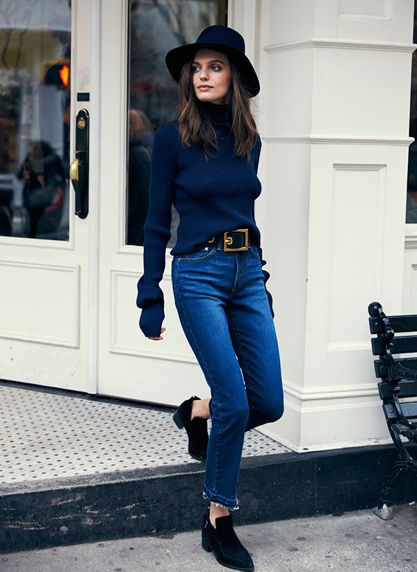 Mulher desce escadaria usando calça jeans de cintura alta cropped flare com tomboy shoes preto, turtleneck azul marinho e chapéu preto
