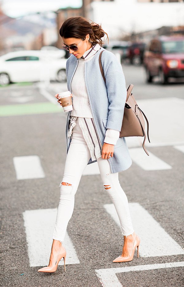 Christine Andrew atravessa a rua posando para foto de street style usando camisa com listras, sueter branco, casaco azul serenity, calça jeans branca skinny destroyed e scarpin nude