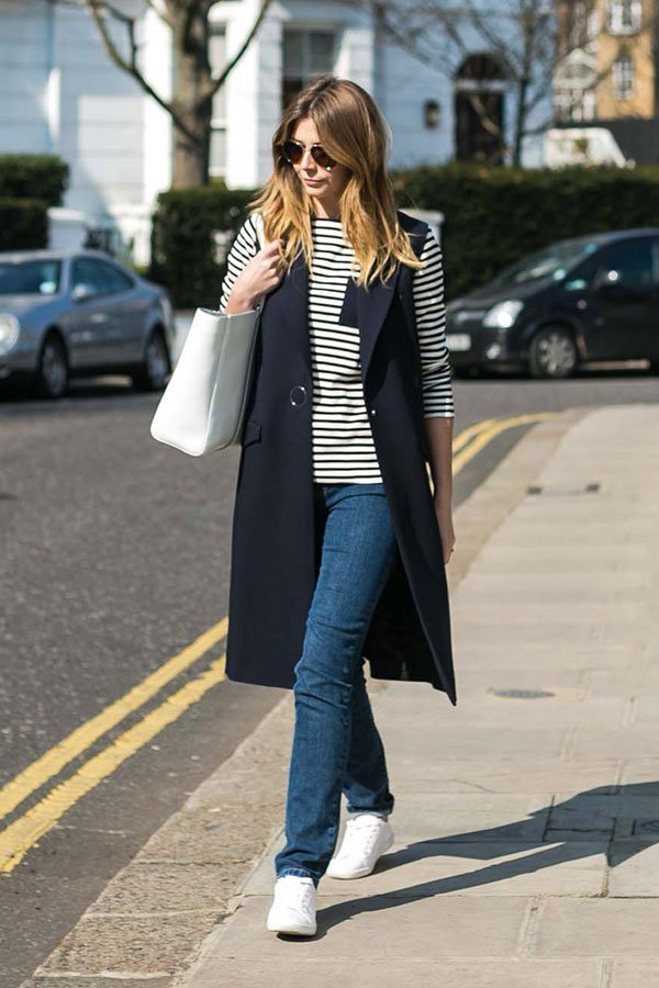 Mulher morena andando nas ruas de Londres vestindo blusa de manga comprida listrada, calça jeans de modelagem reta, colete longo azul marinho, tênis branco, bolsa branca e óculos escuros
