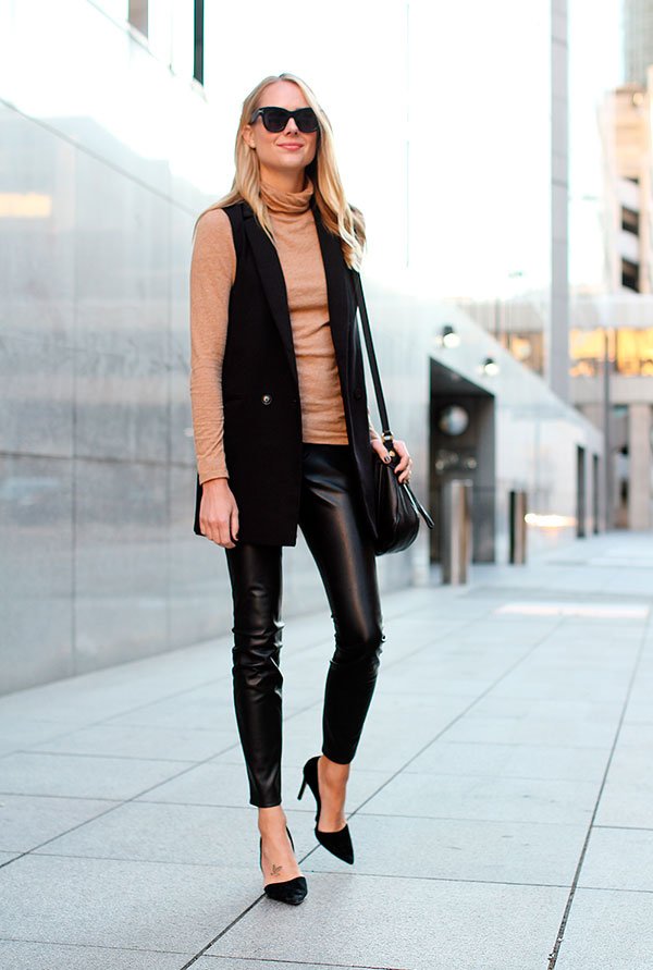 Mulher loira parada na calçada posa para foto vestindo turtleneck caramelo, colete longo preto, calça skinny preta, scarpin preto, óculos escuros e bolsa preta