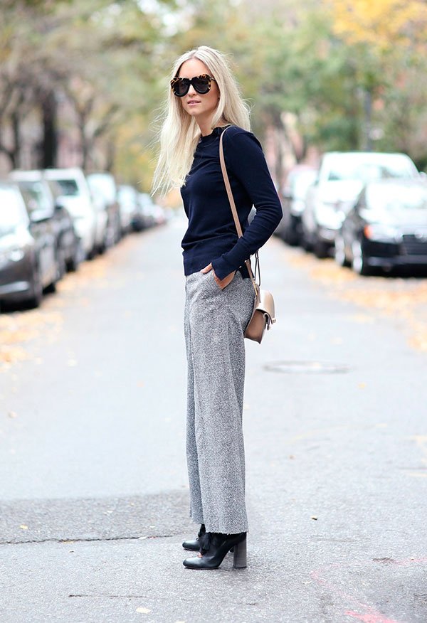 Loira parada no meio da rua posando para foto de street style usando calça cinza cropped, botas, blusa de manga longa, bolsa a tiracolo e óculos escuro