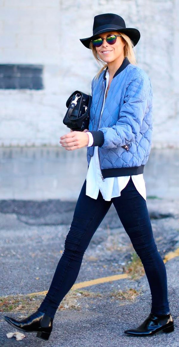 Mulher loira andando na rua usando calça jeans skinny preta, botas de verniz, camisa branca, jaqueta bomber azul serenity, bolsa proenza schouler e chapéu preto