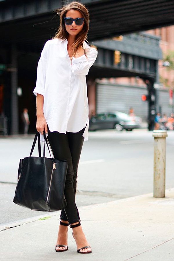 Mulher posa para foto de street style usando camisa branca, calça de couro preta, sandália de tiras, óculos escuros e bolsa saco preta