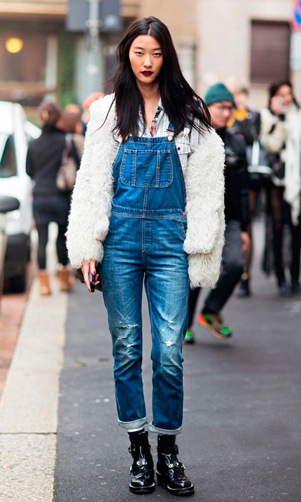 Mulher posa para foto na rua usando camisa jeans clara, jardineira jeans azul, botas de verniz preta, casaco faux fur bege