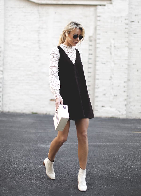 Mulher loira andando na rua posa para foto de street style usando camisa de renda branca, vestido de camurça preto, botas brancas e bolsa baú branca