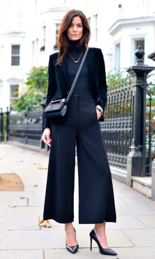 Mulher morena posa para foto na calçada de Londres usando turtleneck preta com blazer de veludo preto por cima, calça pantalona cropped, scarpin preto e bolsa a tiracolo na transversal