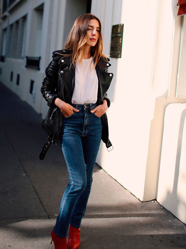 Sarah Nait andando na rua posa para foto usando t-shirt branca, calça jeans de cintura alta, botas vermelhas e  jaqueta de couro preta