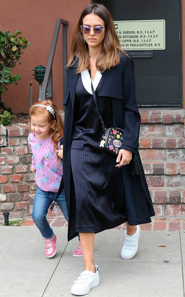 Jessica Alba com a filha usa look com vestido azul marinho com risca de giz, casaco sobretudo azul marinho, óculos gatinho, bolsa com patches e tênis branco