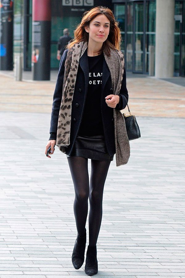 Street style look Alexa Chung com saia mini de couro preta, meia calça, tshirt frase, casaco e lenço onça.