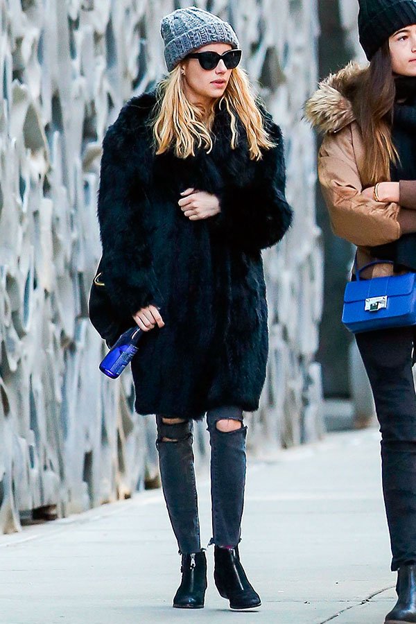 Street style look Emma Roberts usando maxi casaco pêlos preto, calça rasgada, bota cano baixo, gorro cinza e maxi óculos de sol.