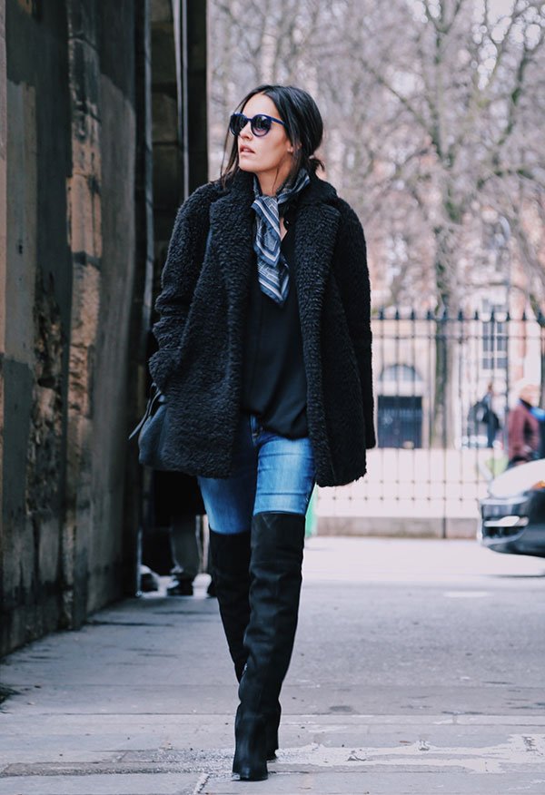 Street style look com calça jeans, bota over the knee, maxi casaco preto pêlos e óculos preto.
