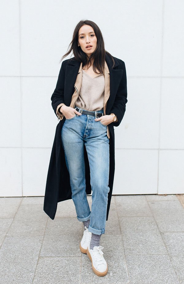 Street style look blusa bege, calça jeans, tênis branco e sobretudo preto.