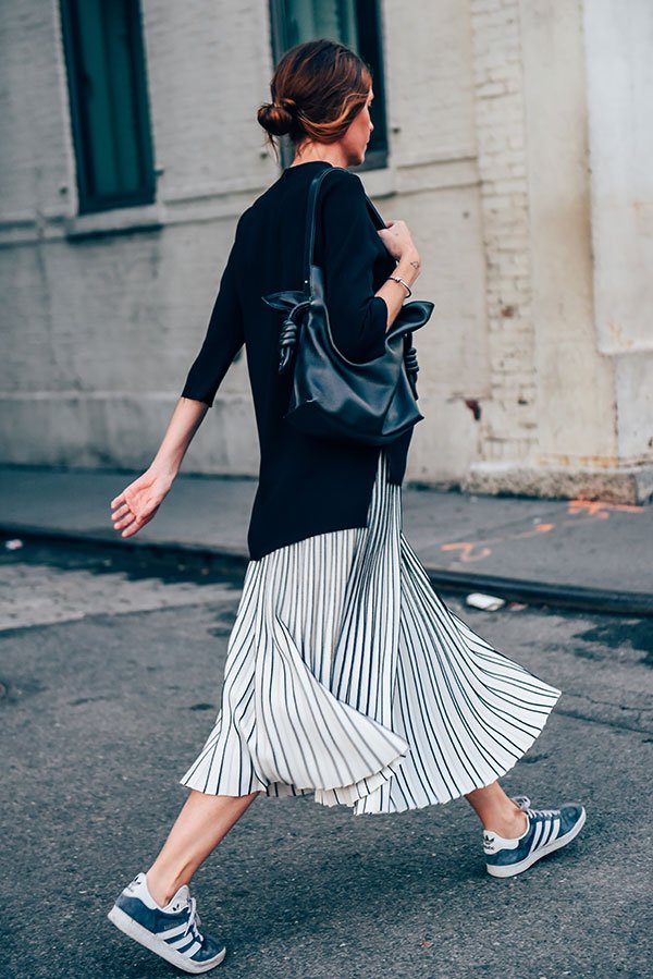 Foto de street style de mulher morena usando look despojado com tricot preto, saia midi de listras preta e branca, bolsa de couro e tênis adidas gazelle azul