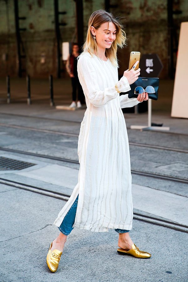 Street style look com maxi vestido branco sobre calça jeans e sapatos dourados.