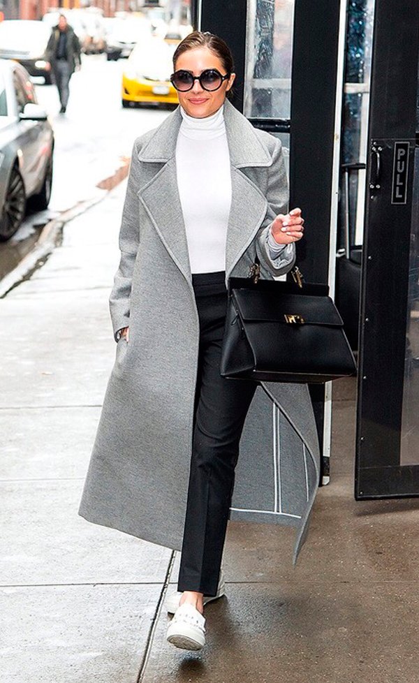 Olivia Culpo usa turtleneck branca, calça de alfaiataria preta, casaco sobretudo cinza, tênis branco e bolsa estruturada preta, finalizando com óculos redondo