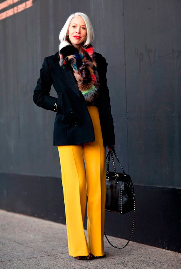 Street style look com calça pantalona amarela, casaco preto e pêlos coloridos.