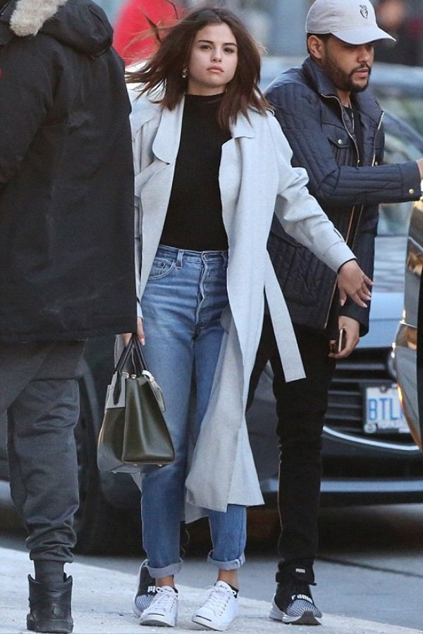 Selena Gomez com casaco longo cinza claro, blusa preta, mom jeans azul claro com a barra dobrada e tênis branco. Segura uma bolsa estruturada verde e preta. Estilo casual urbano. Tendências: minimalismo e camadas.