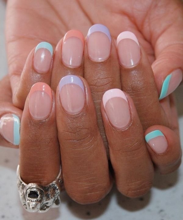 Unha redonda decoradas com esmalte francês em tons pastel, combinando rosa, azul, roxo e verde menta, tendência moderna e estilosa. Acessório destaque: anel de prata em formato de crânio. Ideal para quem busca um visual ousado e elegante.