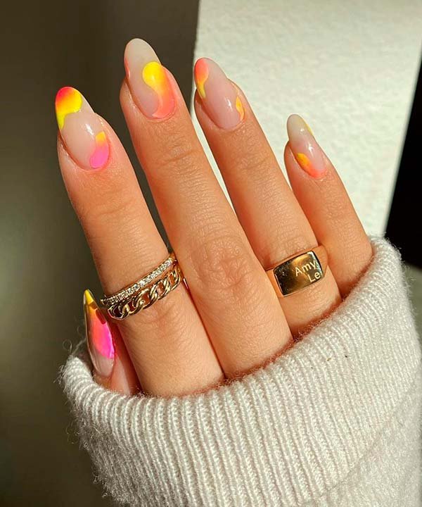 Alongamento de unhas em gel em formato amendoado com nail art gradiente em tons de rosa, amarelo e laranja. A mão está adornada com anéis dourados, incluindo um anel de corrente e um com gravação.