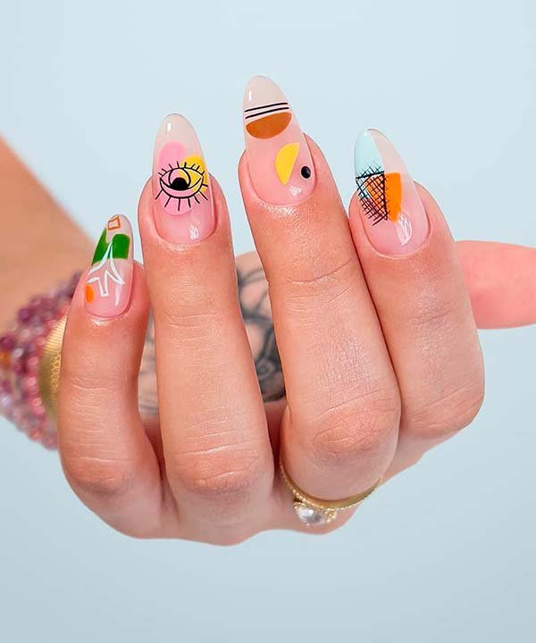 Unhas de fibra de vidro decoradas com nail art abstrato e colorido, seguindo as tendências modernas. Desenhos incluem formas geométricas e artísticas. Anel dourado e pulseiras de miçangas compõem o visual.