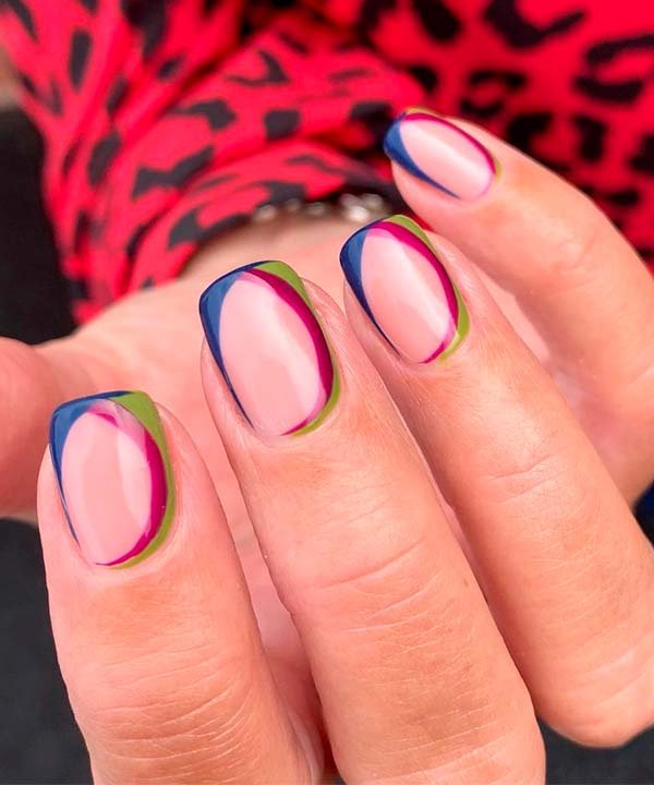 Detalhe de unhas com nail art geométrica em tons de azul, rosa e verde, destacando-se contra uma blusa vermelha com estampa de oncinha. Estilo moderno e vibrante, ideal para quem busca tendências ousadas na moda e beleza.