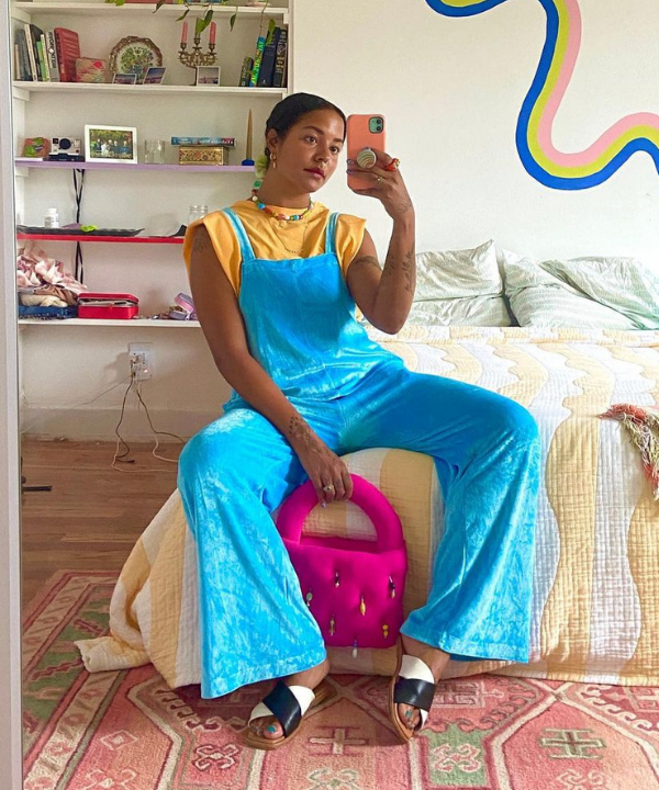Michelle Norris - macacão azul com camiseta amarela e sandália rasteirinha de tiras cruzadas - sandália rasteirinha - Verão 2022 - sentada na cama - https://stealthelook.com.br