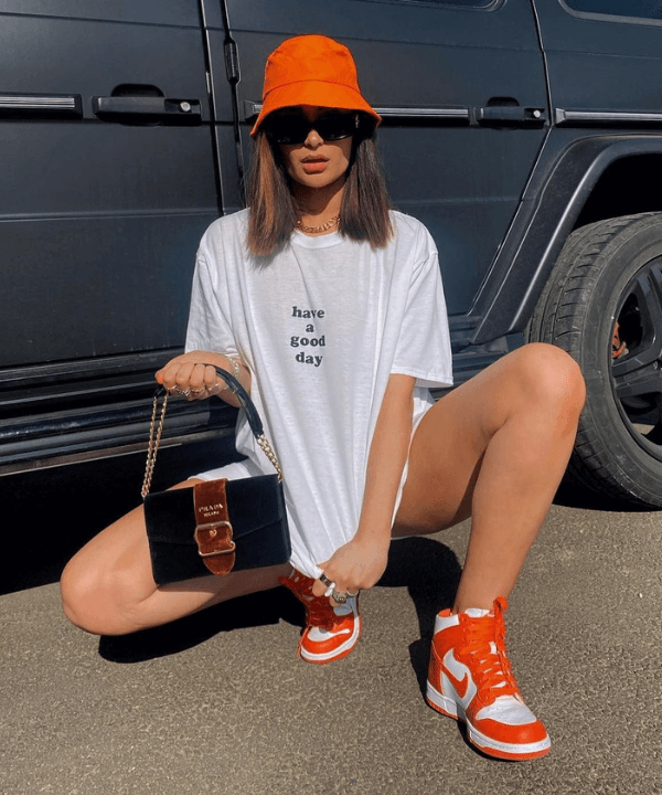 Alicia Roddy - camiseta branca e bucket hat laranja - look com camiseta branca - Verão - sentada na rua - https://stealthelook.com.br