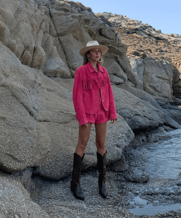 Sofia Coelho - jaqueta de franja rosa e shorts com botas de cowboy - tendência dos anos 70 - verão - em pé perto de pedras - https://stealthelook.com.br