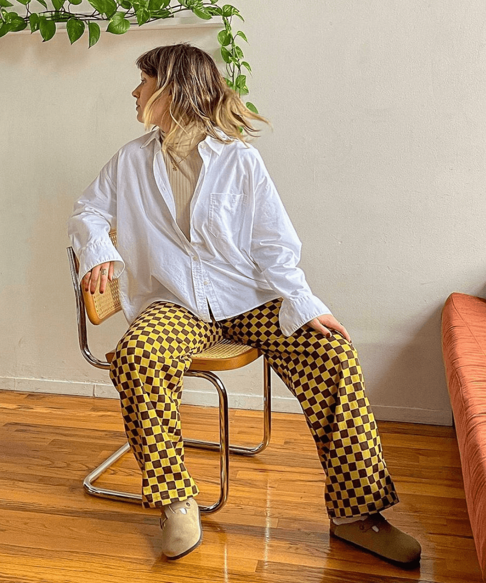 Sophie Wood - calça quadriculada amarela e marrom, camisa branca e blusa de gola alta bege - looks novos - Outono - sentada em uma cadeira olhando para o lado - https://stealthelook.com.br