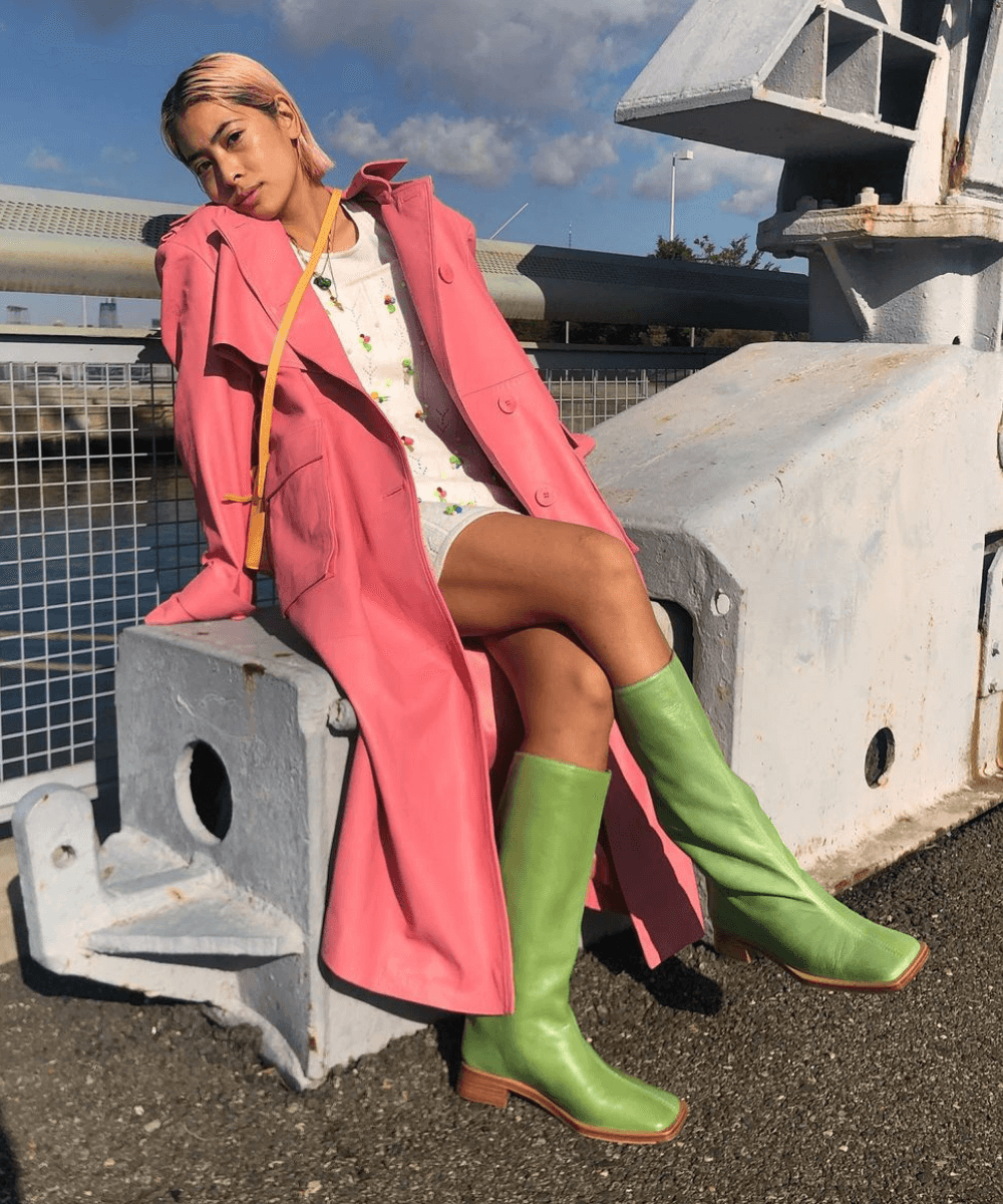 Michelle Li - vestido curto branco, botas verdes de cano longo e trench coat rosa - looks novos - Outono - sentada em uma mureta na rua com as pernas cruzadas - https://stealthelook.com.br