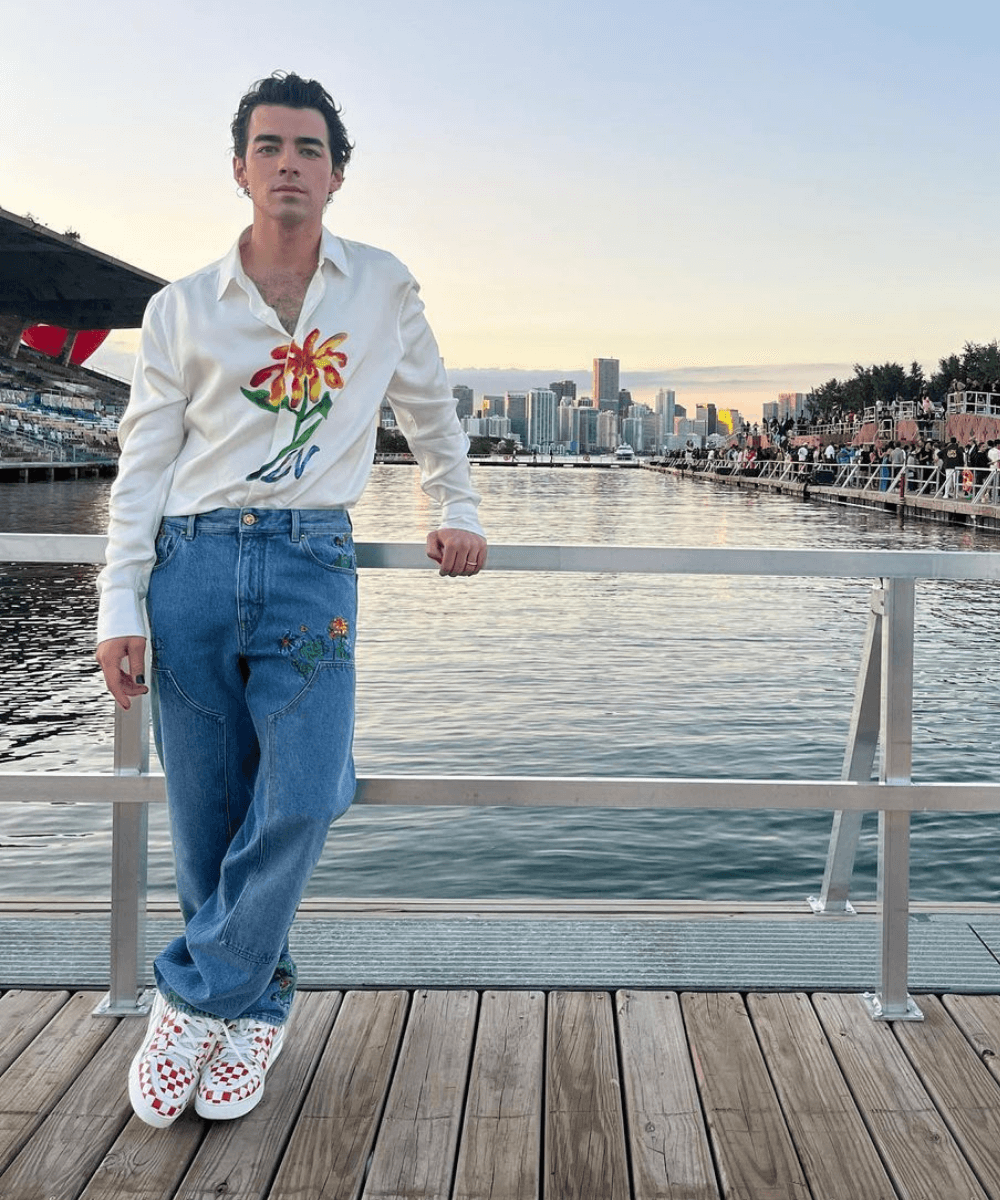 Joe Jonas - calça jeans, tênis quadriculado e cardigan branco com flor - Joe Jonas - Verão - em pé em um pier - https://stealthelook.com.br