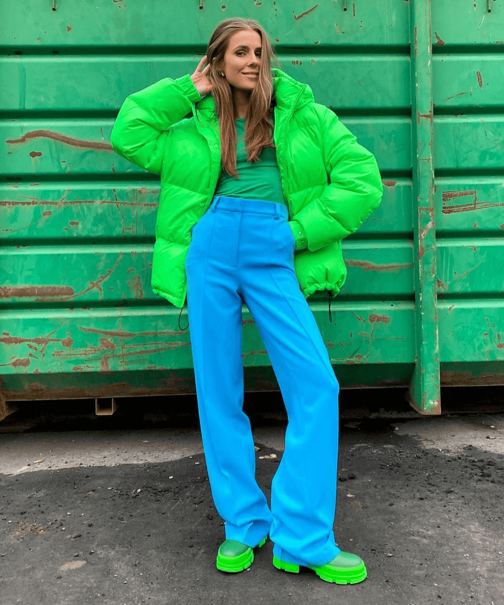 Nina Sandbech - calça azul, jaqueta puffer verde neon e botas - dopamine dressing - Inverno  - em pé na rua em frente a uma parede verde - https://stealthelook.com.br