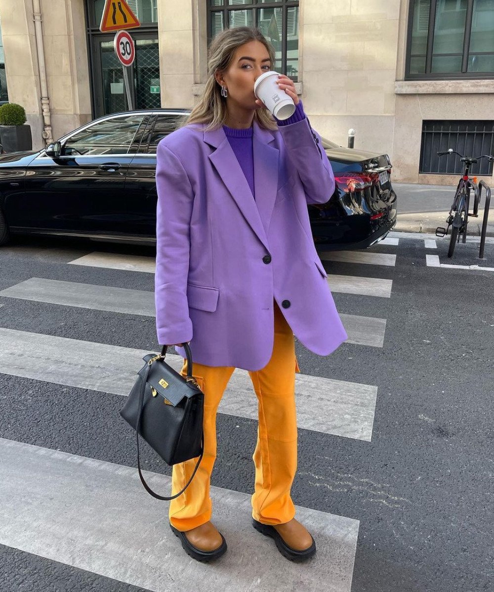 Emili Sindlev - calça laranja, blazer roxo oversized e botas - looks coloridos - Outono - em pé na rua tomando um café - https://stealthelook.com.br