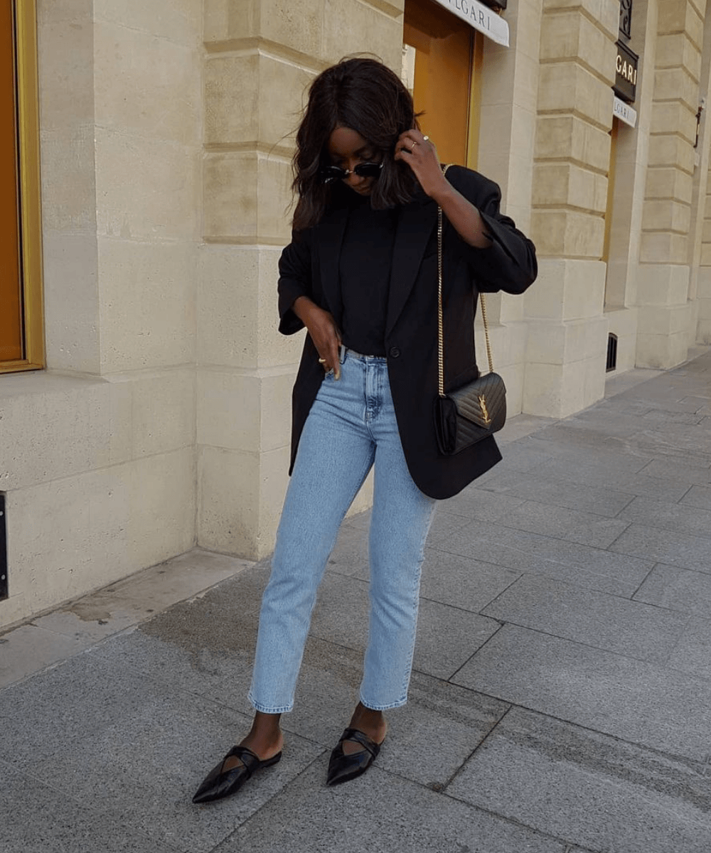 Aïda Badji Sané - calça jeans reta, blusa preta, blazer preto e mules de bico fino - sapatos confortáveis - Primavera - em pé na rua olhando para o chão com a mão no cabelo - https://stealthelook.com.br