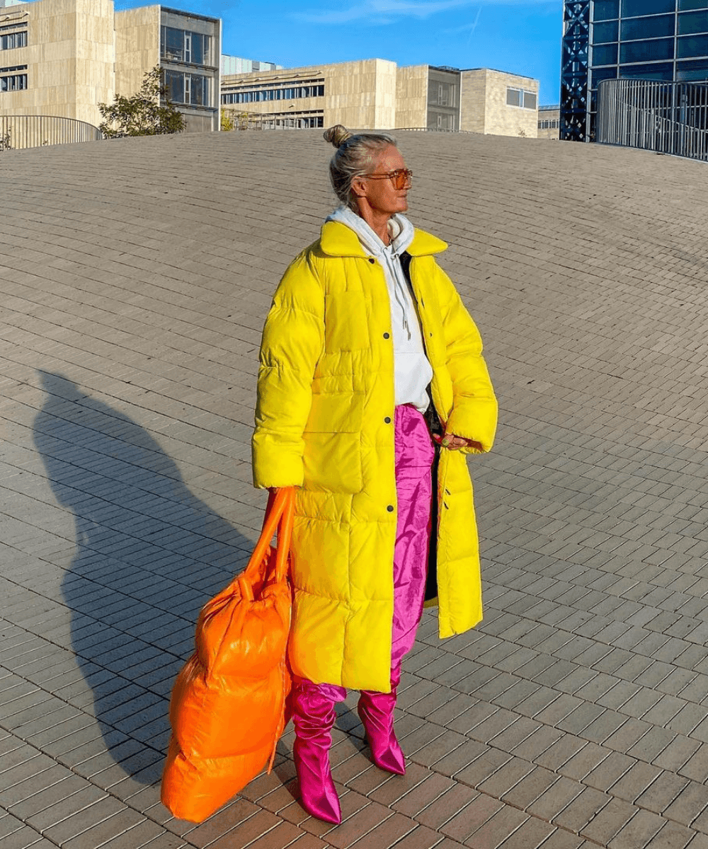 Mette | @msorrig - calça rosa, bota rosa e casaco puffer longo amarelo - tendência de moda - Inverno  - em pé na rua segurando uma bolsa laranja acolchoada grande - https://stealthelook.com.br