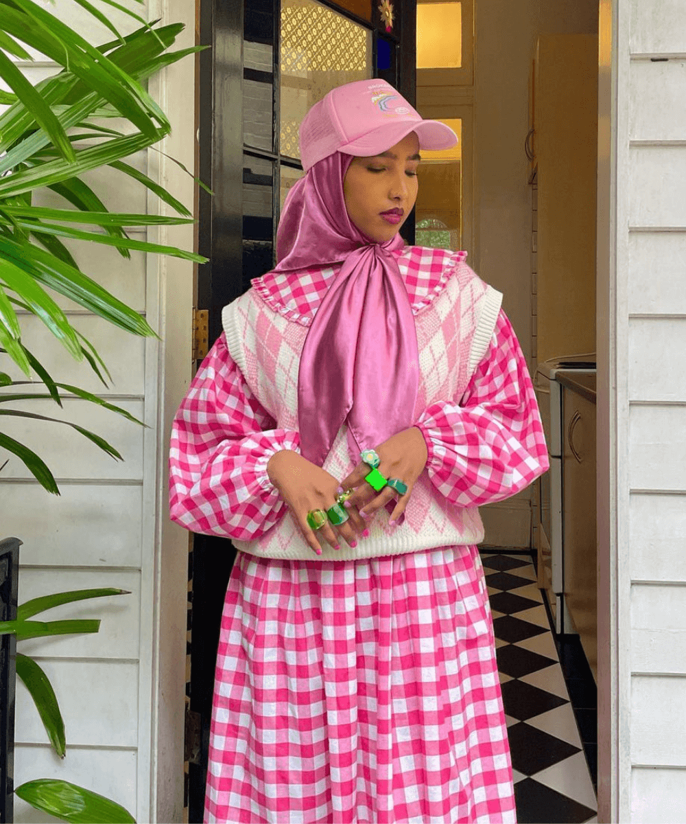 Basra Ajeh | @itsbasra - vestido xadrez, colete estampado e boné rosa com anéis verdes no dedo - outono 2022 - Outono - em pé na frente de uma porta branca - https://stealthelook.com.br
