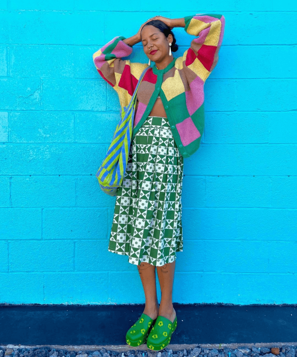 Michelle Norris - cardigan de tricô quadriculado com saia floral e clogs verde - sapatos polêmicos - Inverno 2022 - em frente a uma parede - https://stealthelook.com.br