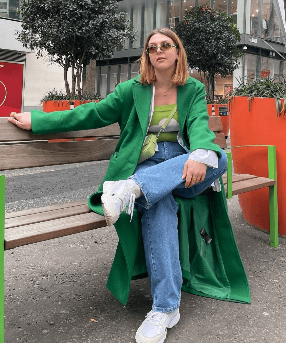 Madison | @Madisoneley - calça jeans, tênis, trench coat verde e blusa estampada - looks novos - Outono - sentada em uma cadeira - https://stealthelook.com.br