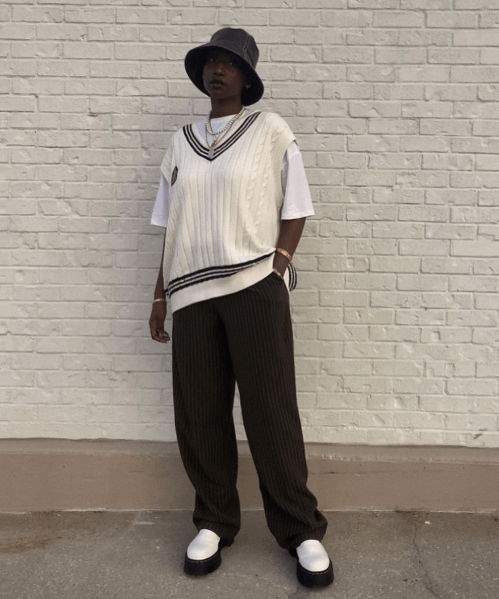 Khadija | @kingkhadija - calça preta, t-shirt oversized branca e colete de tricô - looks novos - Outono - em pé na rua usando um bucket hat preto - https://stealthelook.com.br