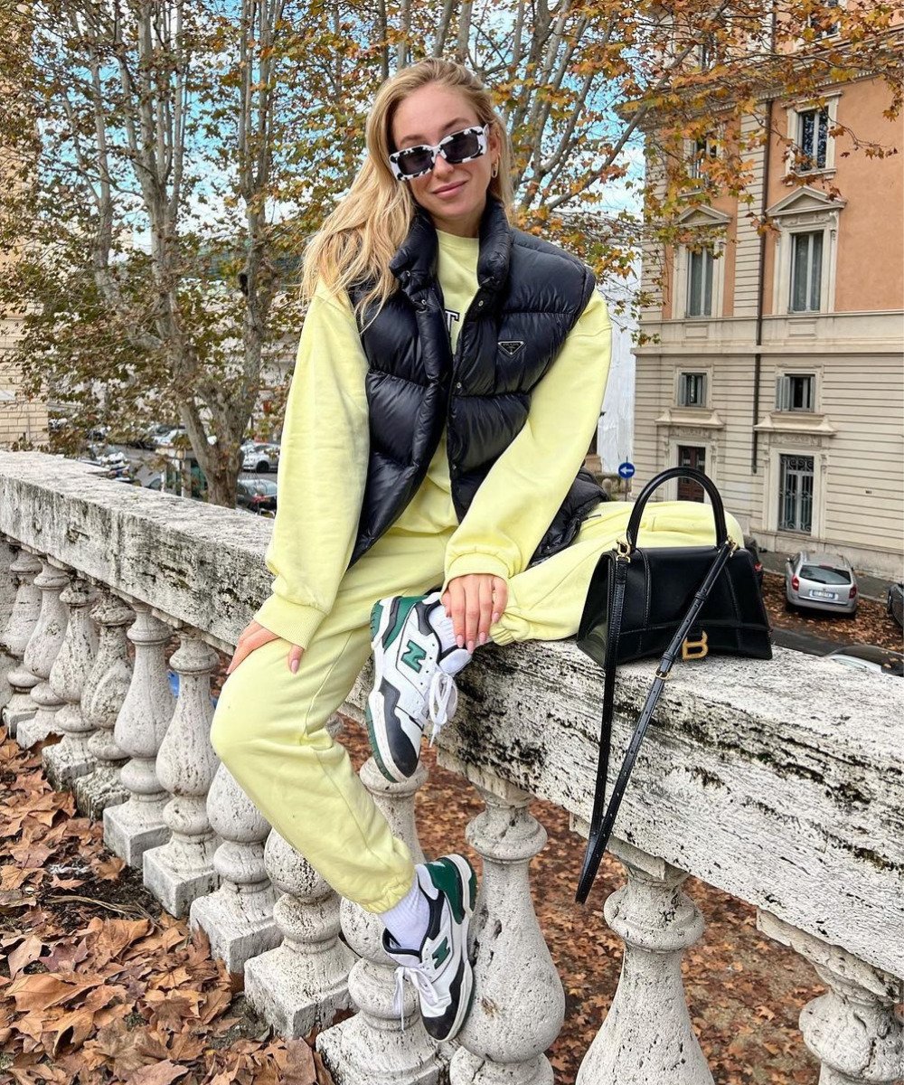Sonia Lyson - conjunto de moletom amarelo, tênis e casaco puffer preto - cores tendência - Inverno  - sentada em uma mureta com a perna esquerda dobrada - https://stealthelook.com.br
