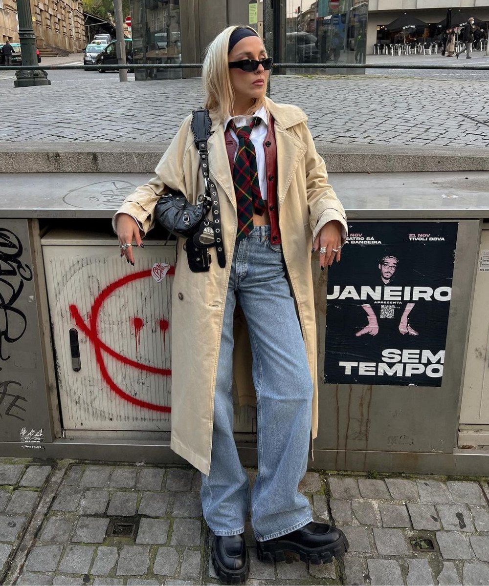 Sofia Coelho - calça jeans lookse, coturno preto, camisa, colete e trench coat - tendências de inverno - Inverno  - em pé na rua na frente de uma grade de metal com posters - https://stealthelook.com.br