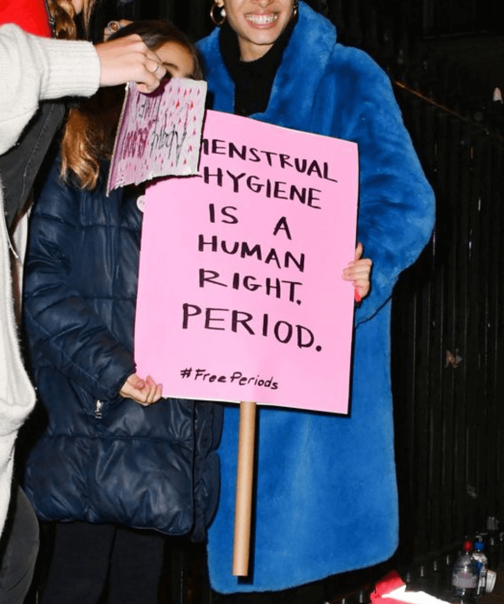 Adwoa Aboah - casaco azul longo - Dia Internacional da Higiene Menstrual - Inverno  - em pé na rua segurando um cartaz rosa escrito 
