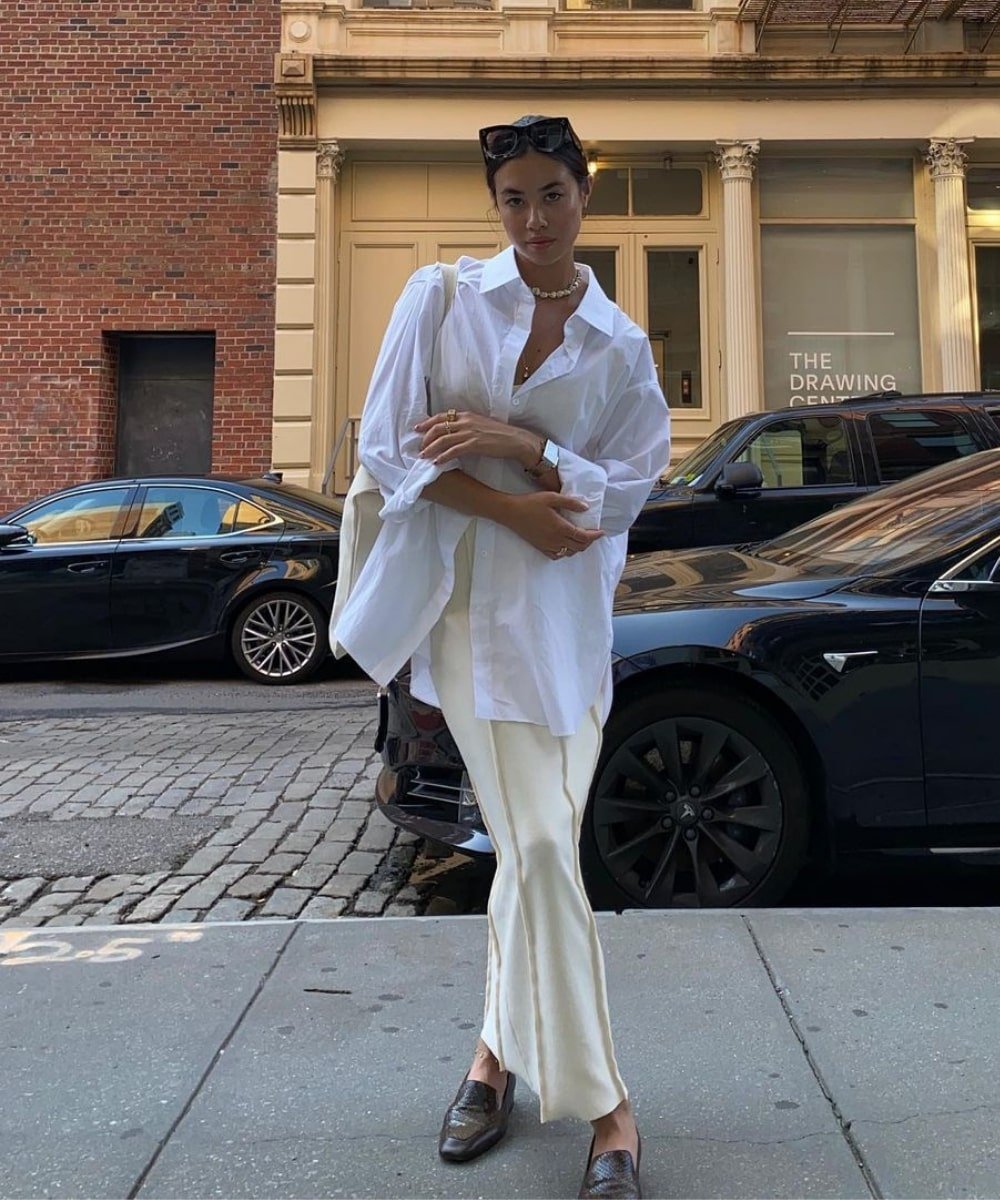 Mulher usando camisa oversized branca, camisa off-white e calça branca, combinados com mocassins escuros e colar de pérolas. Ela segura uma bolsa branca grande e usa óculos de sol no topo da cabeça, representando estilo casual e sofisticado.