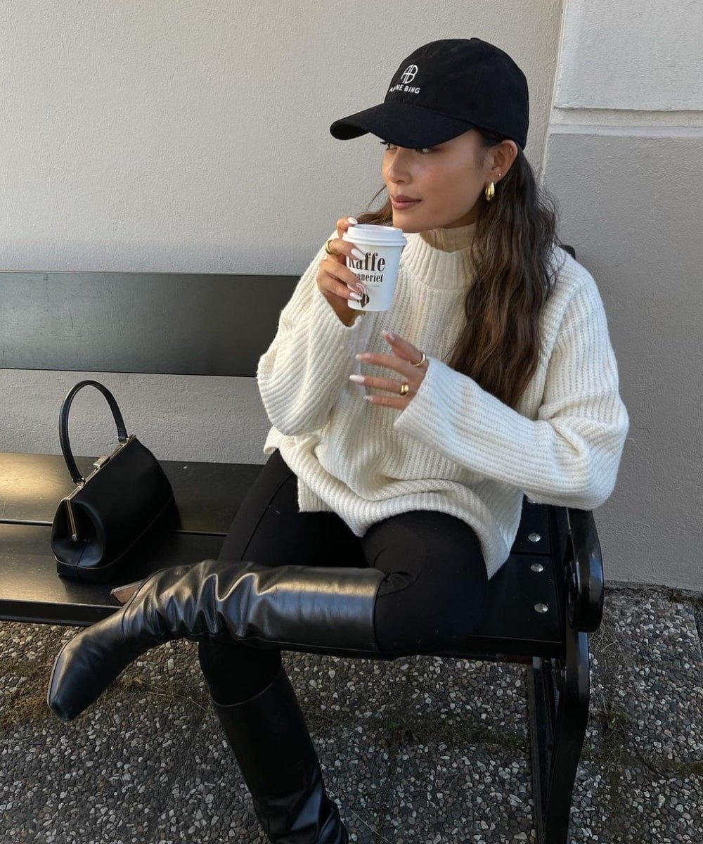 Tsutsumi Hoang - calça legging preta, bota de cano longo, boné e tricô branco - looks novos - Inverno  - sentada em um banco com a perna cruzada - https://stealthelook.com.br